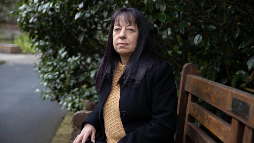 A woman with dark hair sits on a park bench and looks seriously at the camera. 