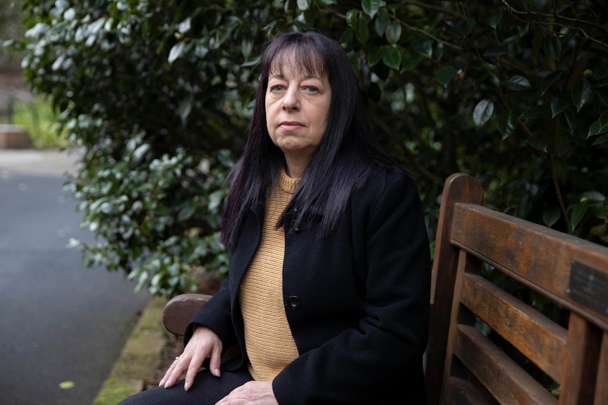 A woman with dark hair sits on a park bench and looks seriously at the camera. 