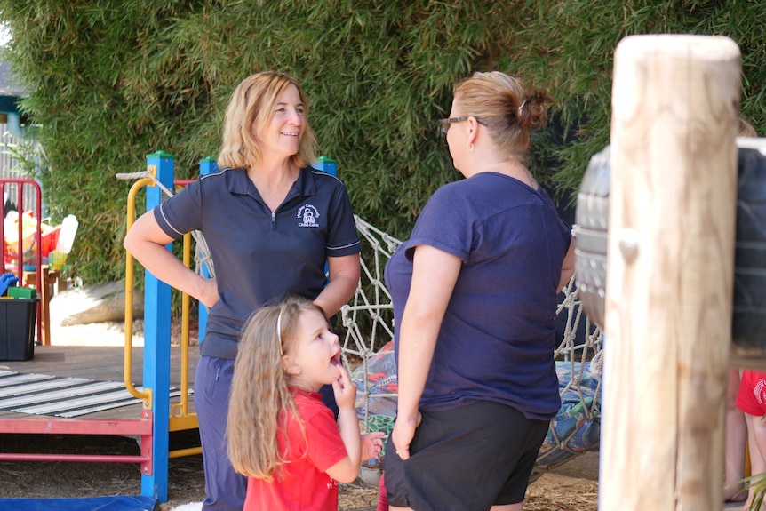 two women working at a childcare centre stand with a young girl, talking