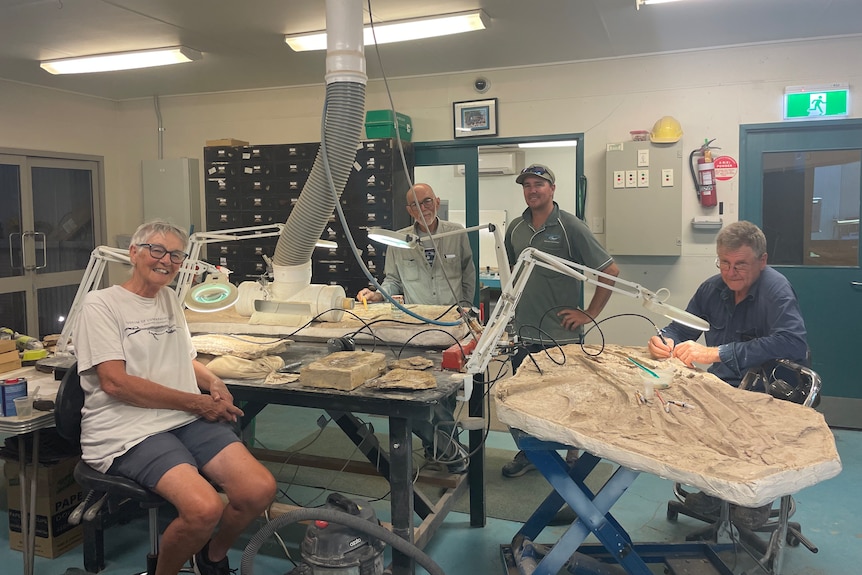 Three men and one women stand and sit in a fossil preparation laboratory.