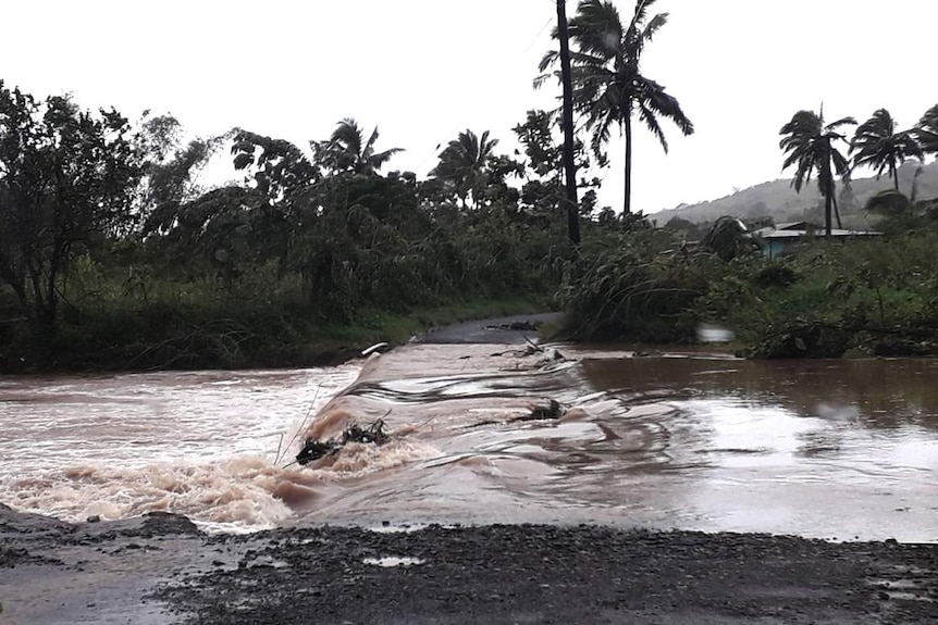 You see water cascading over a shallow bridge on an overcast day with palm trees pushed by heavy winds in the distance.