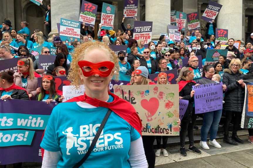 A woman in a teal t-shirt standing in front of a crowd, wearing a red mask.