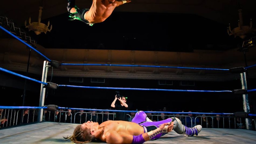 A wrestler in the ring, while another wrestler is flying midair.