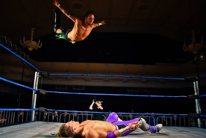 A wrestler in the ring, while another wrestler is flying midair.