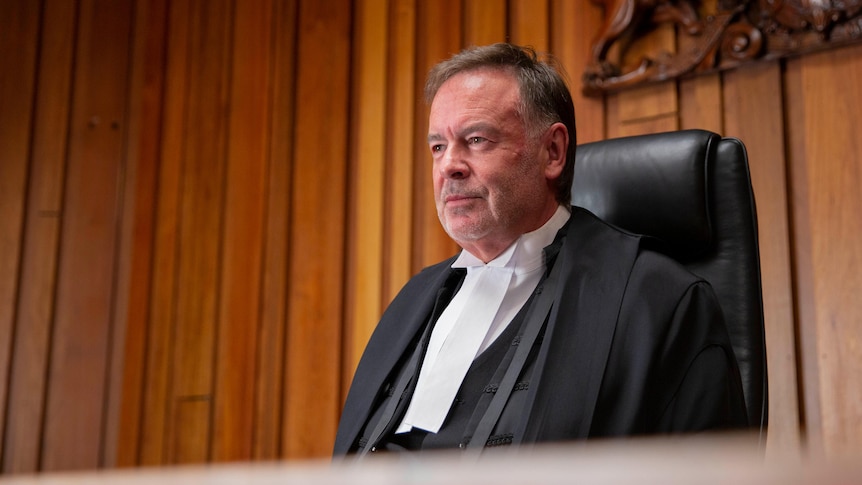A middle aged man in a black legal gown sits in a wood-lined court room. 