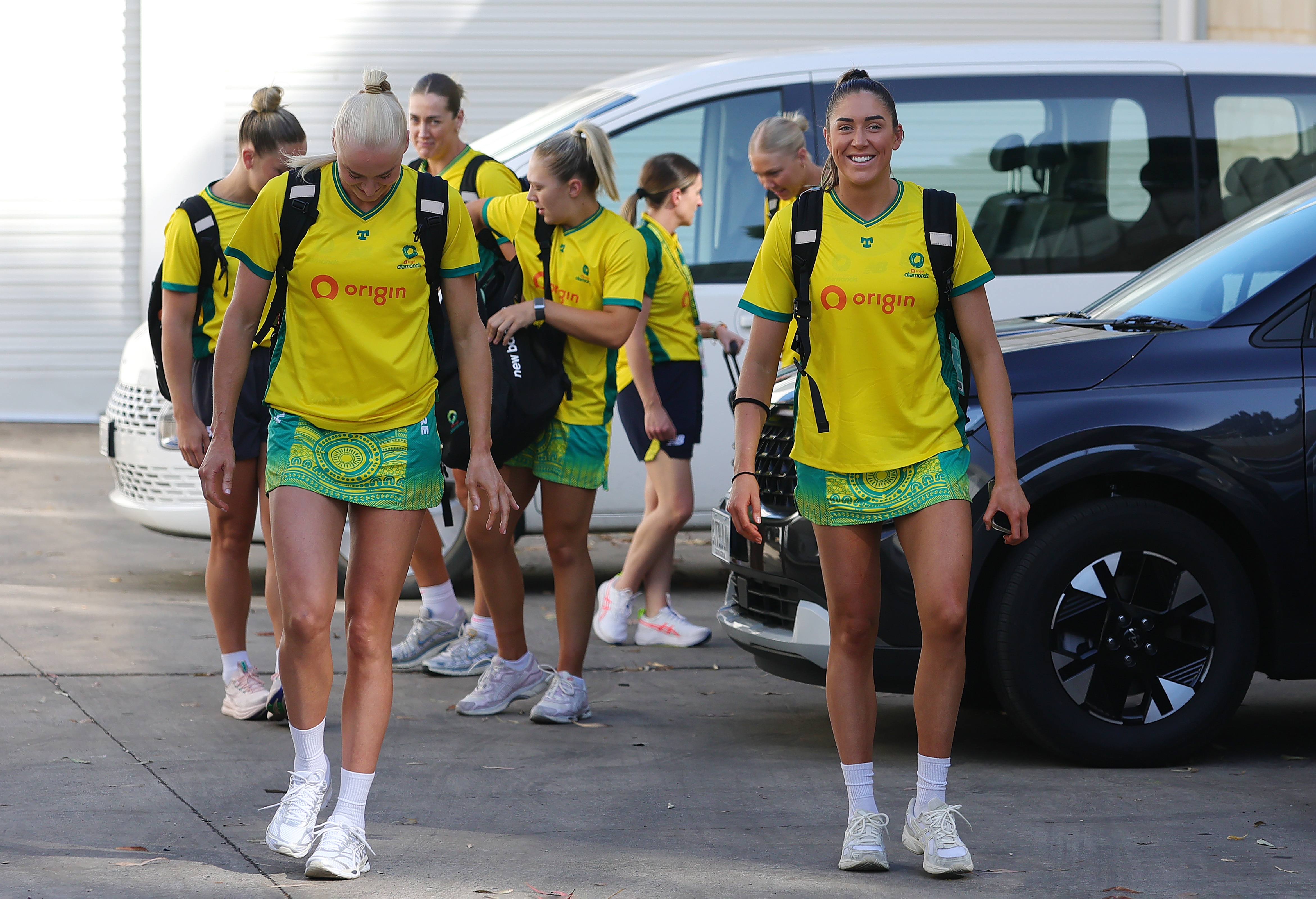 A group of tall netballers in yellow shirts and green skirts walk together near cars.