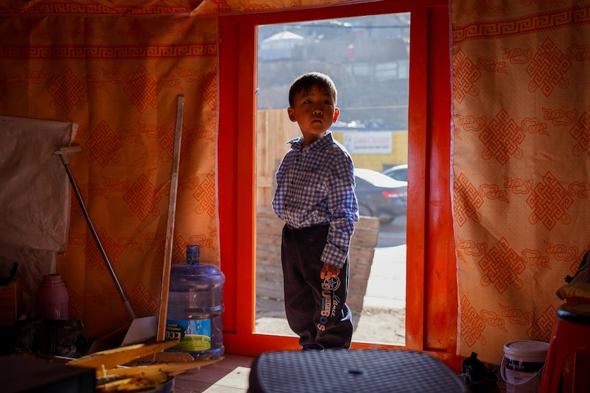 A little boy in blue and white checked shirt stands in the open doorway of a tent-like home, with orange walls