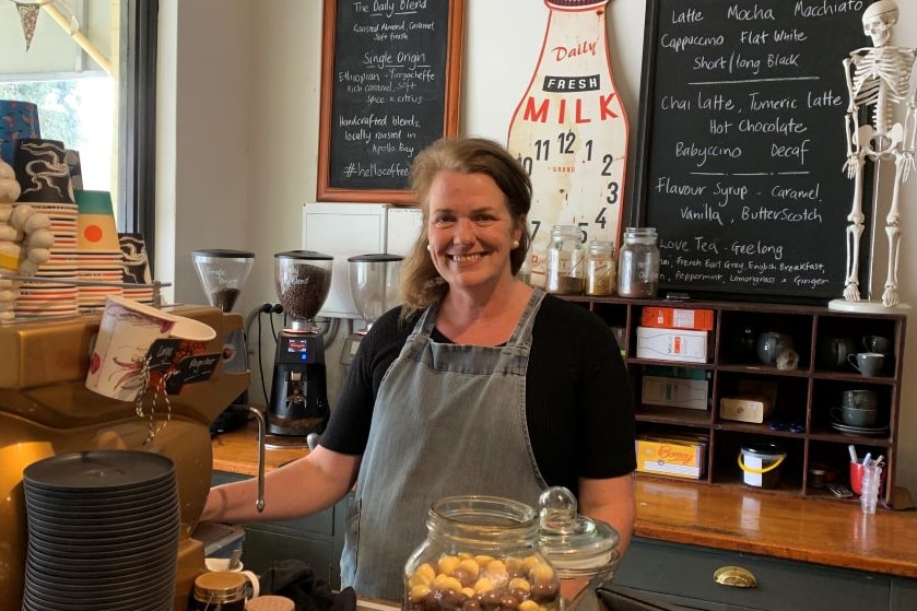 A woman with shoulder length brown hair stands behind a coffee machine and the crowded counter at a country general store.