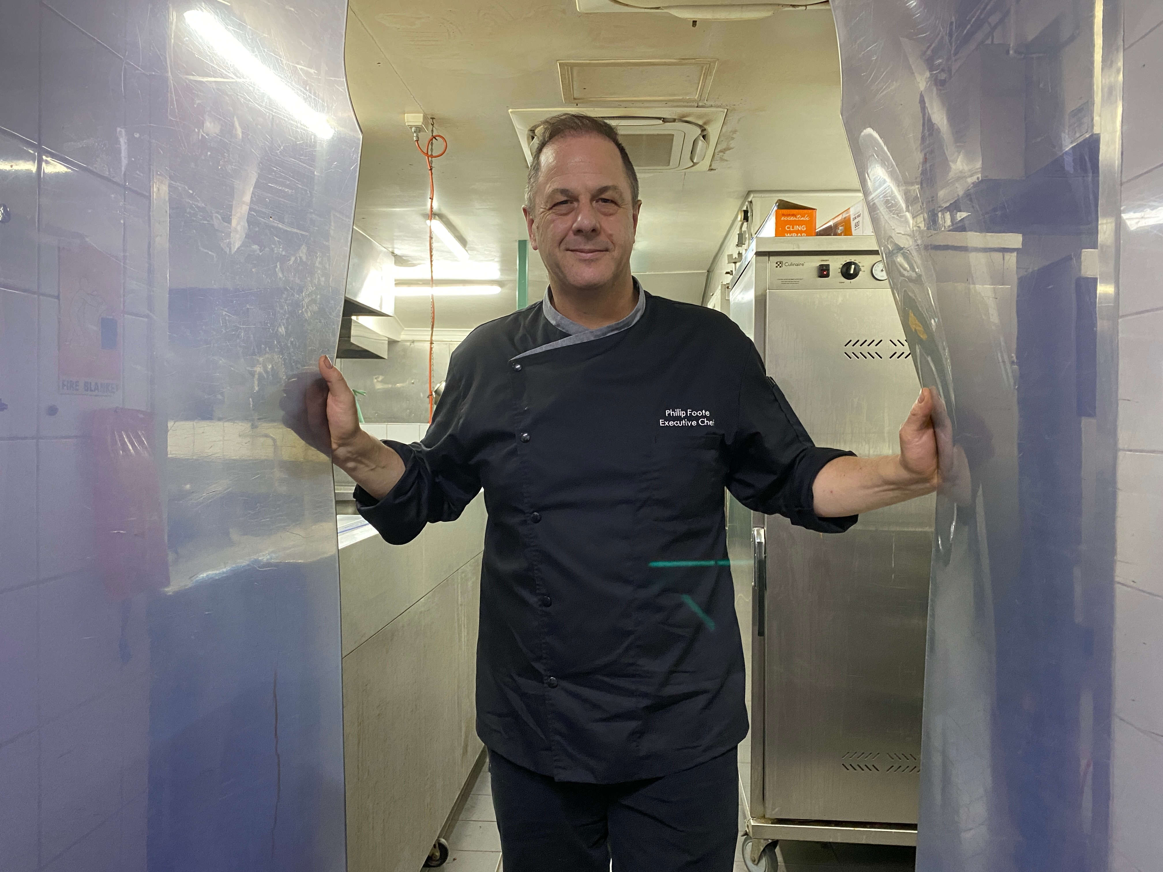A chef wearing black clothes holds open two plastic double doors and stands in the middle of them.