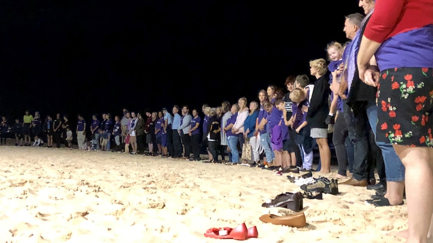 Dozens of people standing in a semi circle on the beach at night with a pair of shoes at their feet.