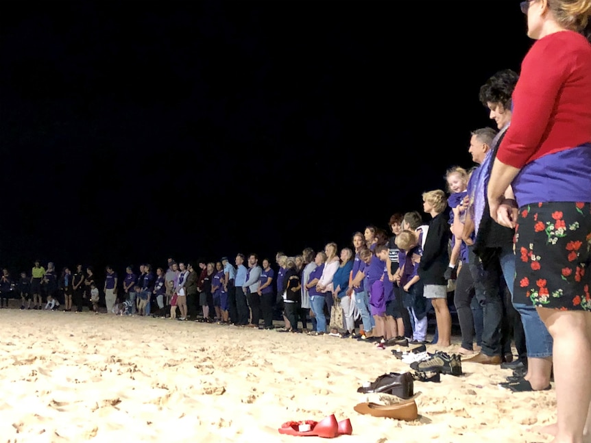Dozens of people standing in a semi circle on the beach at night with a pair of shoes at their feet.