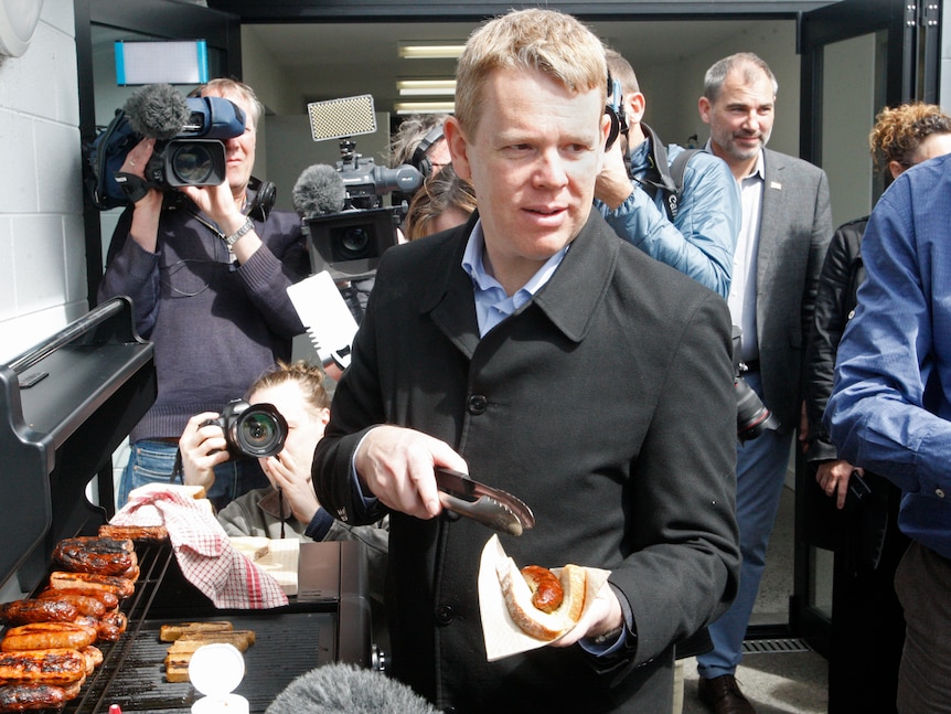 Chris mans a sausage sizzle, holding a sausage in bread. 