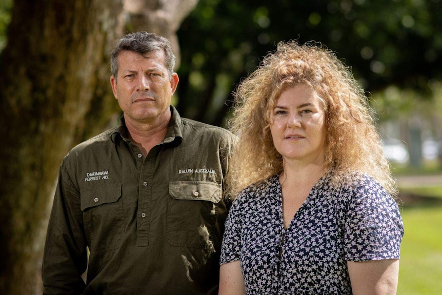 A man with short, greying hair and a woman with long, blonde, frizzy hair stand outdoors, looking serious.