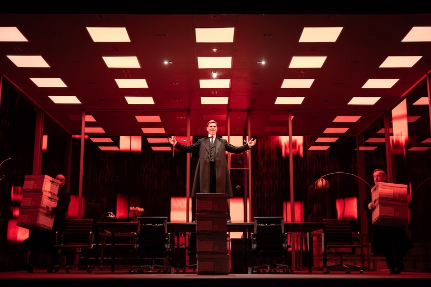 A scene from a stage featuring a male actor standing on a table in a red-lit office