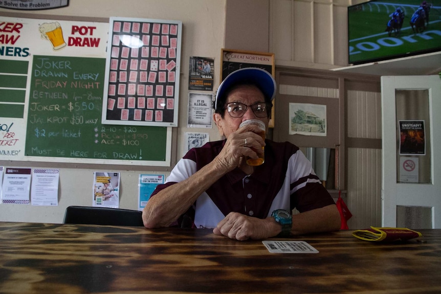 An elderly woman in a blue visor sits at the bar in an outback pub and sips a beer.