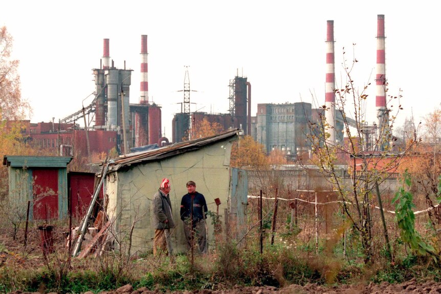 Two men stand outside a shack with pollution stacks in the background 