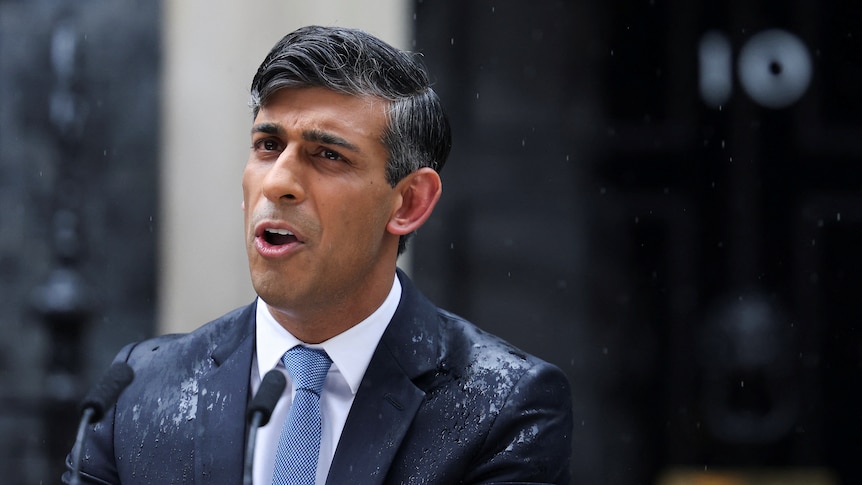 Rishi Sunak stands in a blue suit speaking behind a lectern in the rain in the street