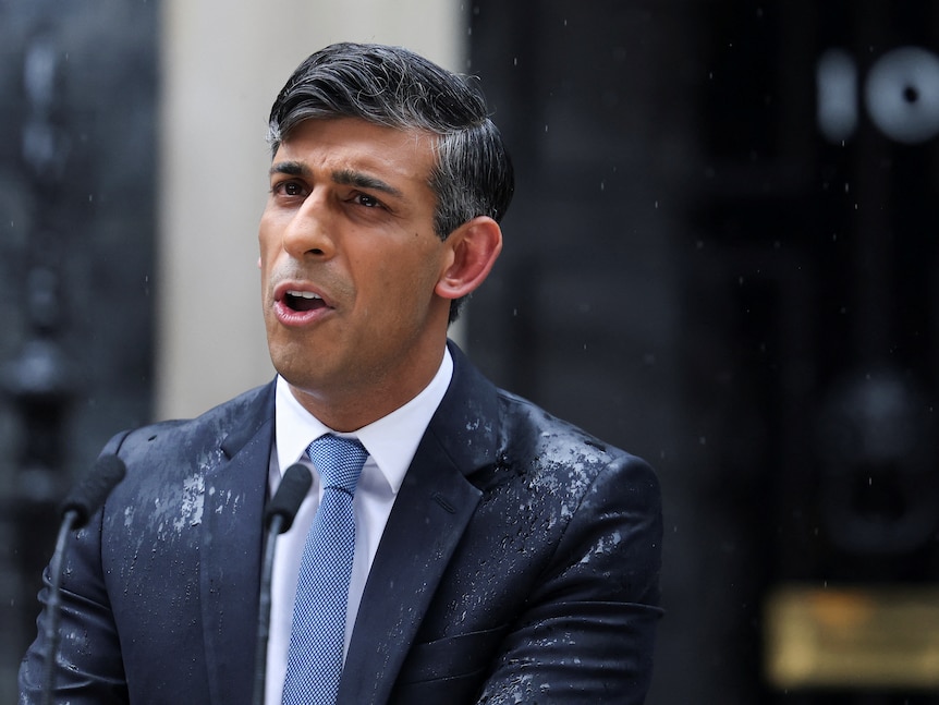 Rishi Sunak stands in a blue suit speaking behind a lectern in the rain in the street