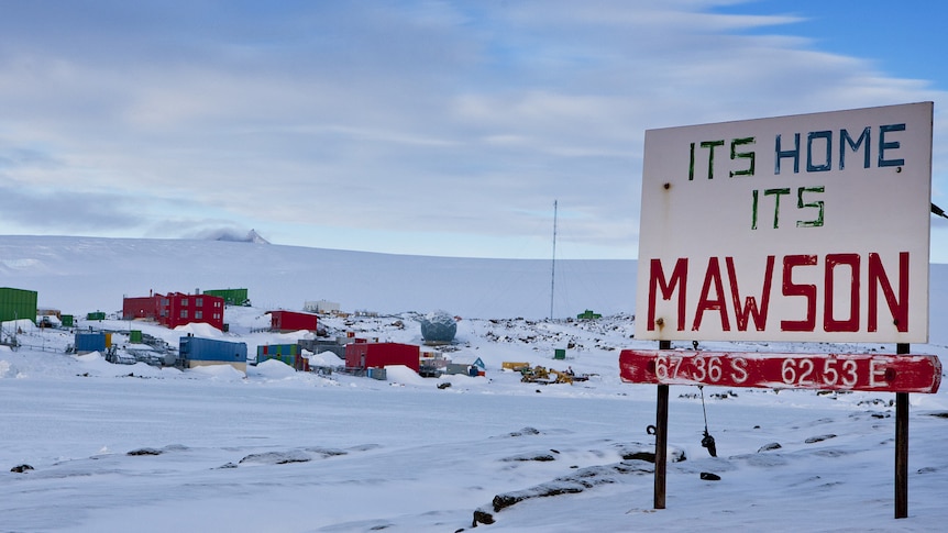 A cluster of coloured buildings in the snow with a sign in the foreground saying "it's home, it's Mawson'. 