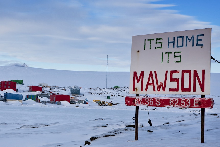 A cluster of coloured buildings in the snow with a sign in the foreground saying "it's home, it's Mawson'. 