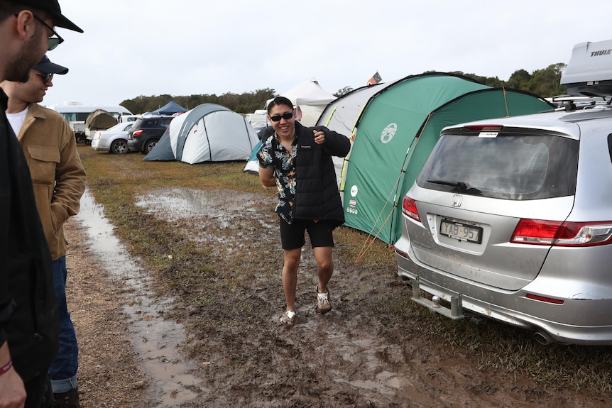 a man puts on a jacket as he walks near tens and cars at a camping ground