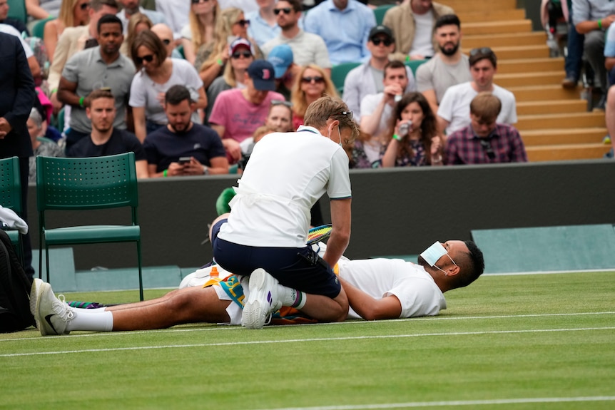 A masked Nick Kyrgios lies on the court at Wimbledon as a medic checks him for an abdominal injury.