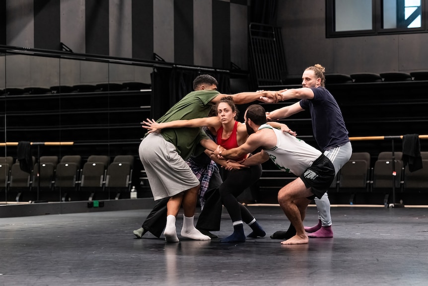 Five First Nations dancers in casual exercise clothes and socks are mid-movement in a grey rehearsal room space.