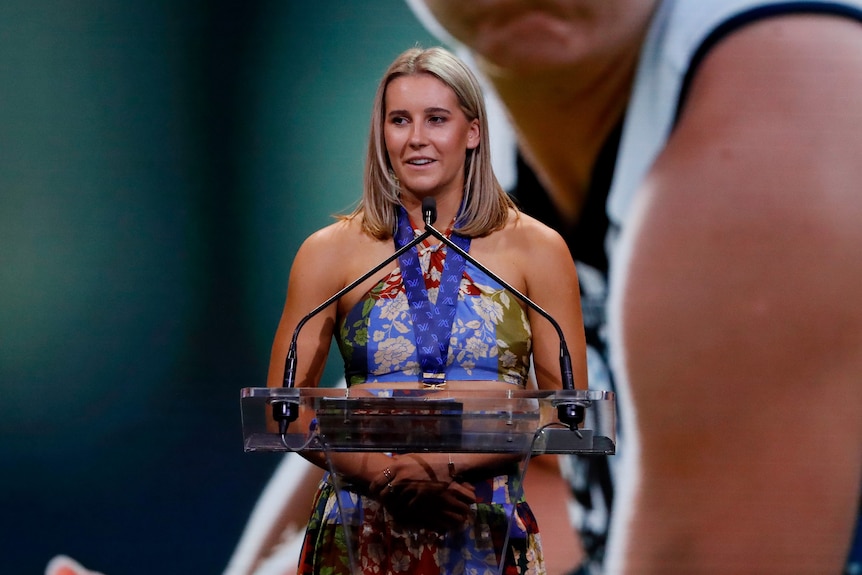 Mimi Hill speaks at a podium wearing a floral dress and a medal with blue ribbon