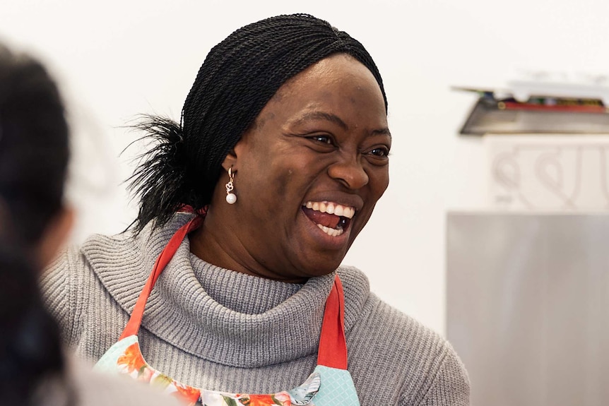 A head and shoulders shot of a laughing woman with black hair wearing a grey jumper and an apron.