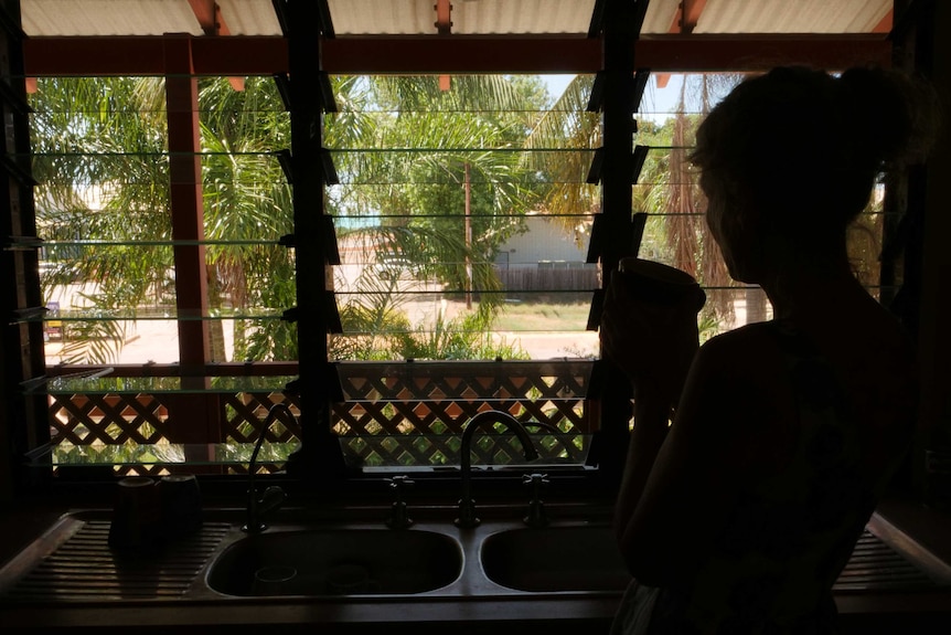 Silhouette of a woman standing at kitchen sink and looking out of louvre windows to palm trees