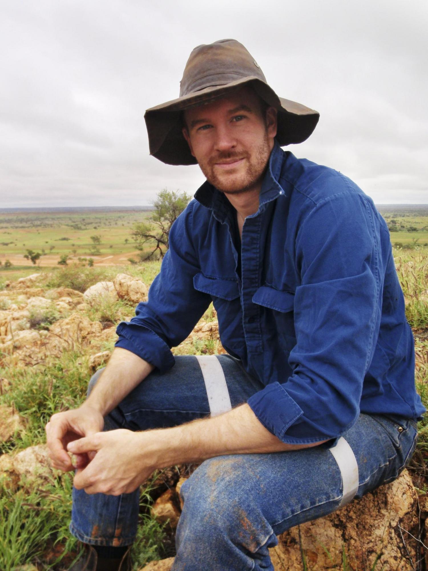 Scientist Dr Thomas Newsome sitting on a rock in the outback wearing a dark blue shirt and brown hat.