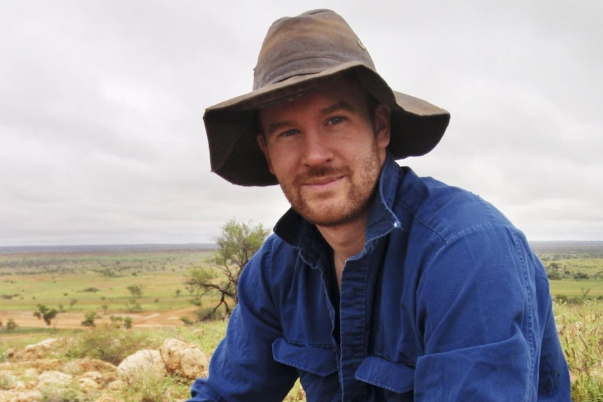 Scientist Dr Thomas Newsome sitting on a rock in the outback wearing a dark blue shirt and brown hat.
