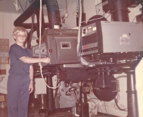 A young man standing behind a movie projector.