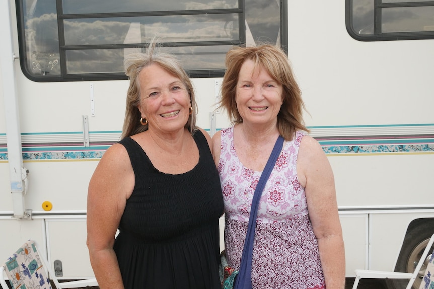 A woman in a black singlet stands beside a woman in a floral singlet, with a van visible behind them.