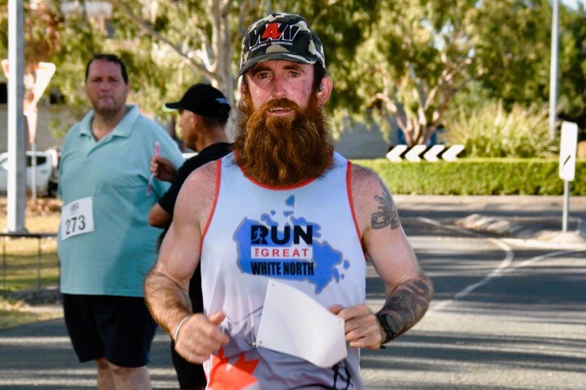 A man with a brown, ginger beard, running and wearing an army style cap and white running singlet.