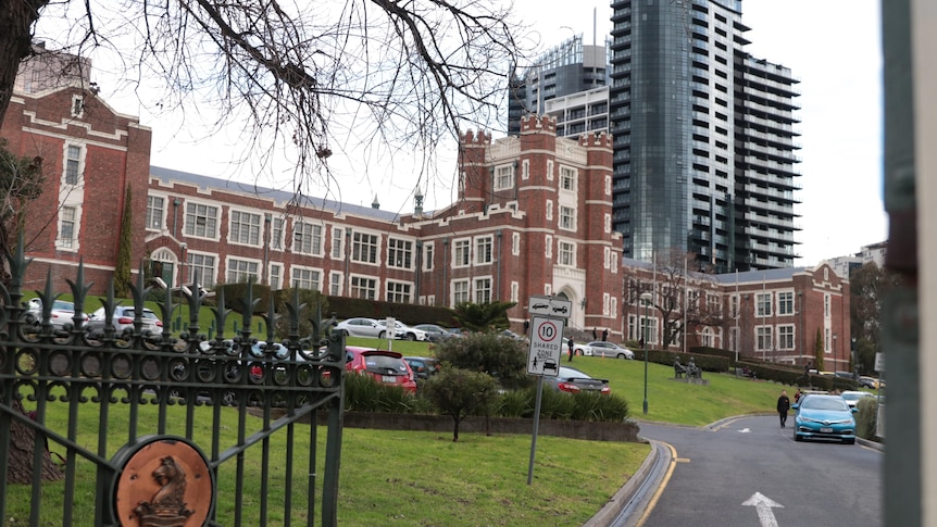 A view of a red brick school building with a gate, driveway and cars in the foreground.