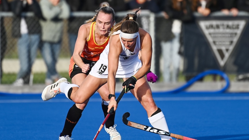 A New Zealand female hockey player tries to control the ball while under pressure from an Australian opponent.