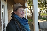 A woman in an Akubra hat stands on a balcony smiles and looks out at a garden 