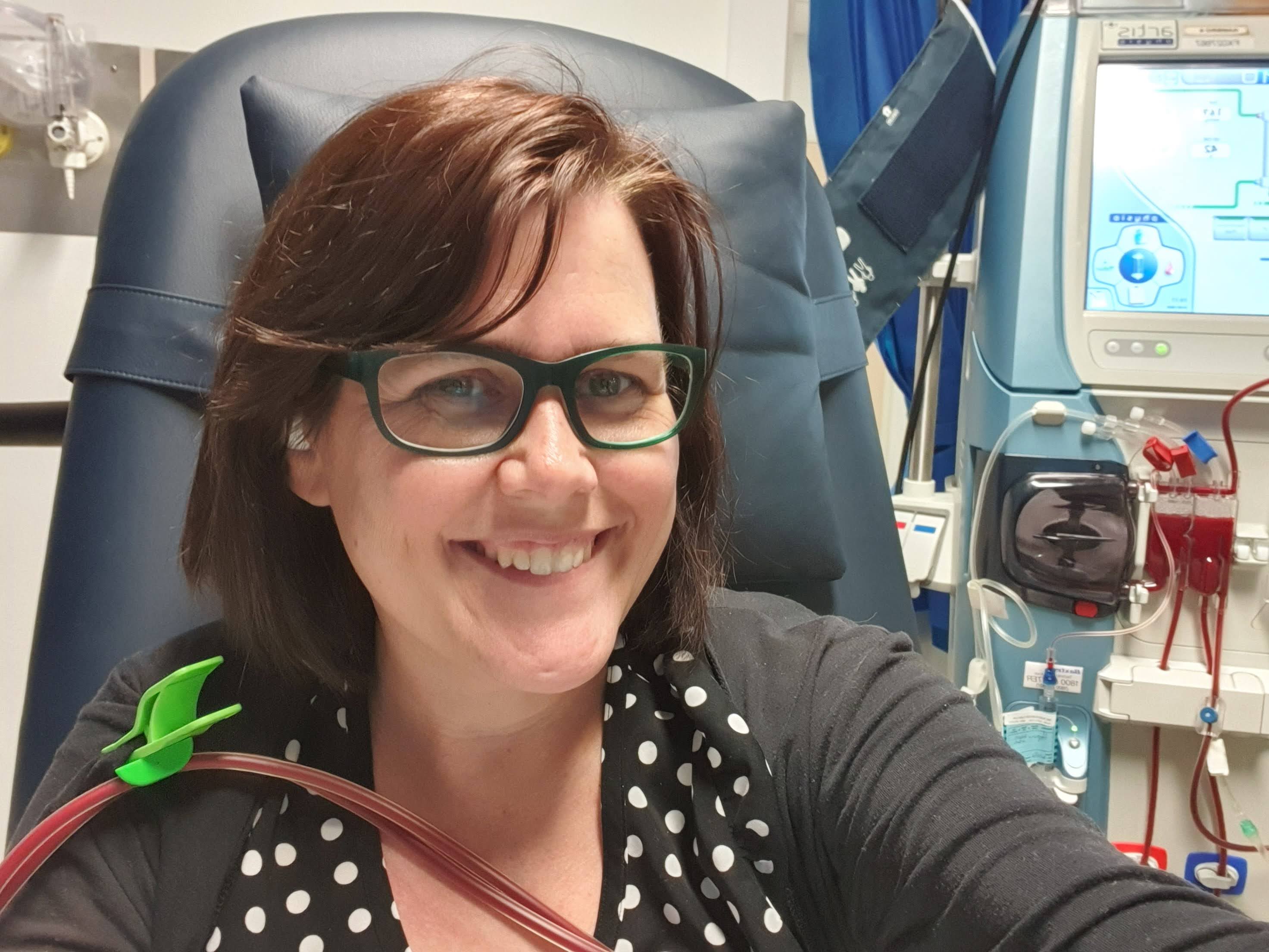 A woman smiling and sitting in a hospital chair having dialysis, the machine sits behind her. 