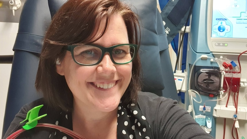 A woman smiling and sitting in a hospital chair having dialysis, the machine sits behind her. 