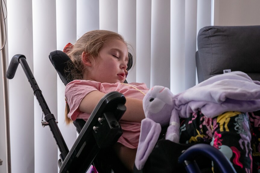 A young girl sits in a wheelchair. Her eyes are closed.