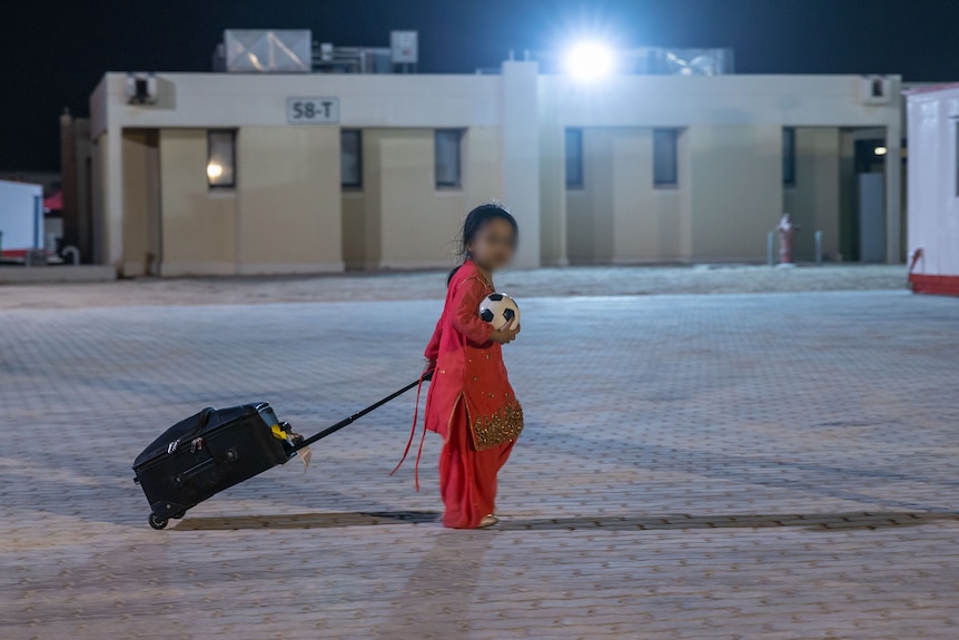 A little girl drags a suitcase on wheels along a military camp.