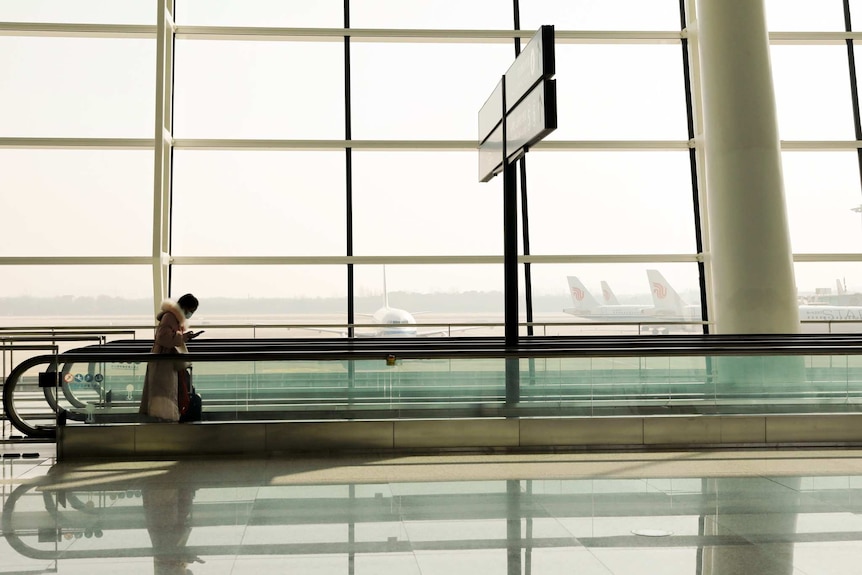 A person wearing a long fur coat and mask looks down at their phone as a plan sits on tarmac in the background.