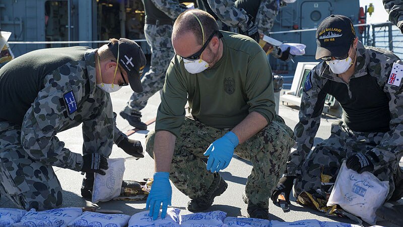 Three men in navy uniforms inspect plastic packages while wearing latex gloves.