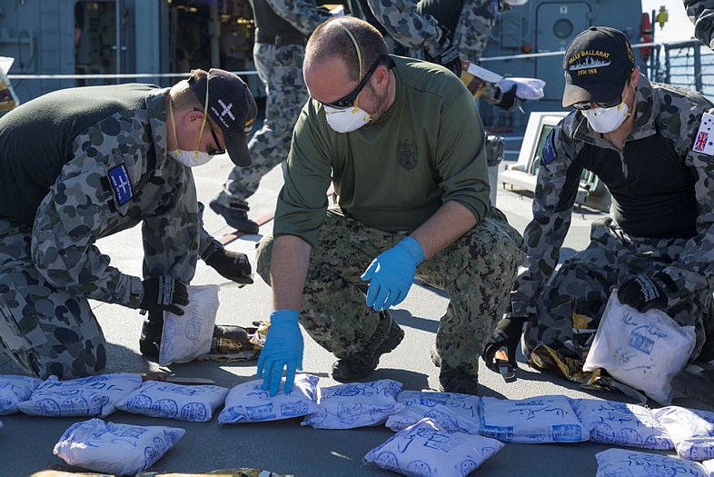 Three men in navy uniforms inspect plastic packages while wearing latex gloves.