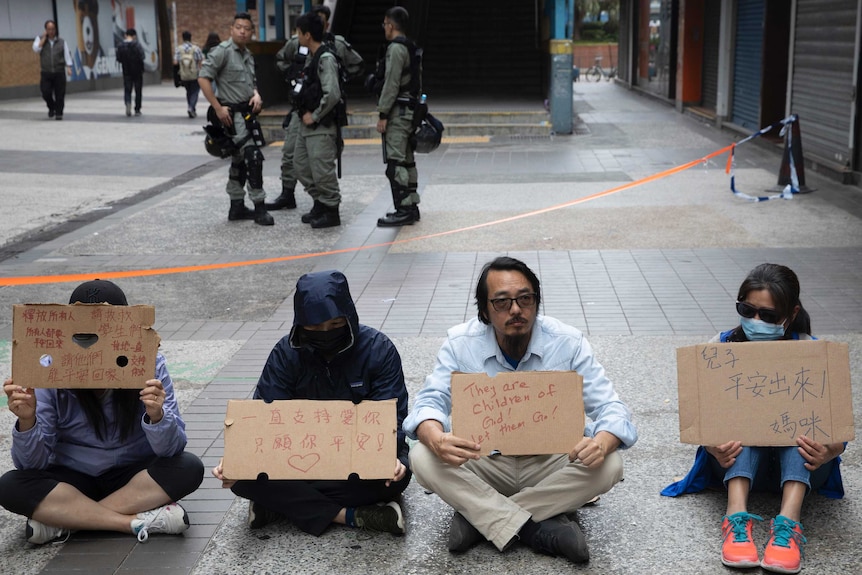 Four people sit with their backs to police holding placard saying they are children of god, let them go.