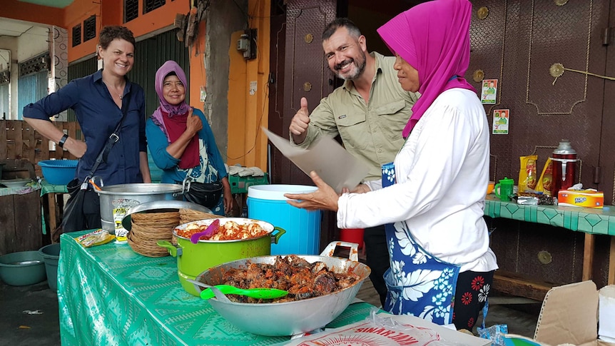 Barker with female stall holders and cameraman giving thumbs up as food served from pots.