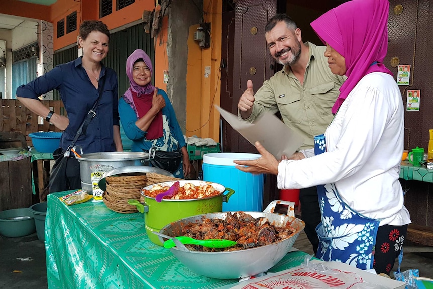 Barker with female stall holders and cameraman giving thumbs up as food served from pots.