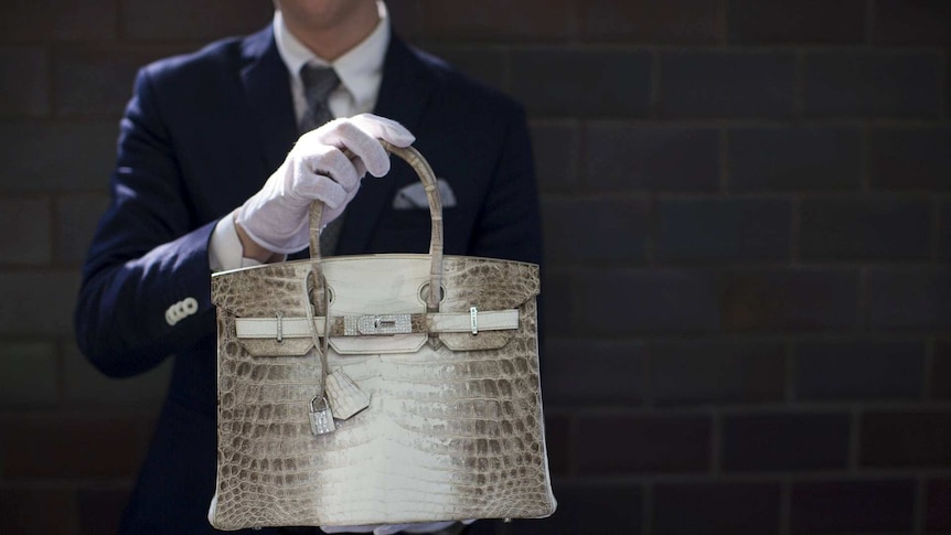 a crocodile skin handbag being held by a man in a suit and white gloves.