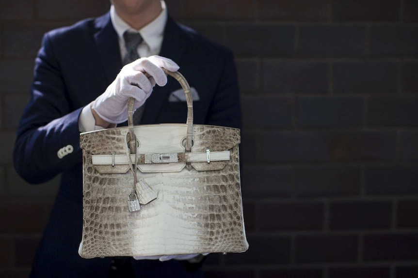 a crocodile skin handbag being held by a man in a suit and white gloves.